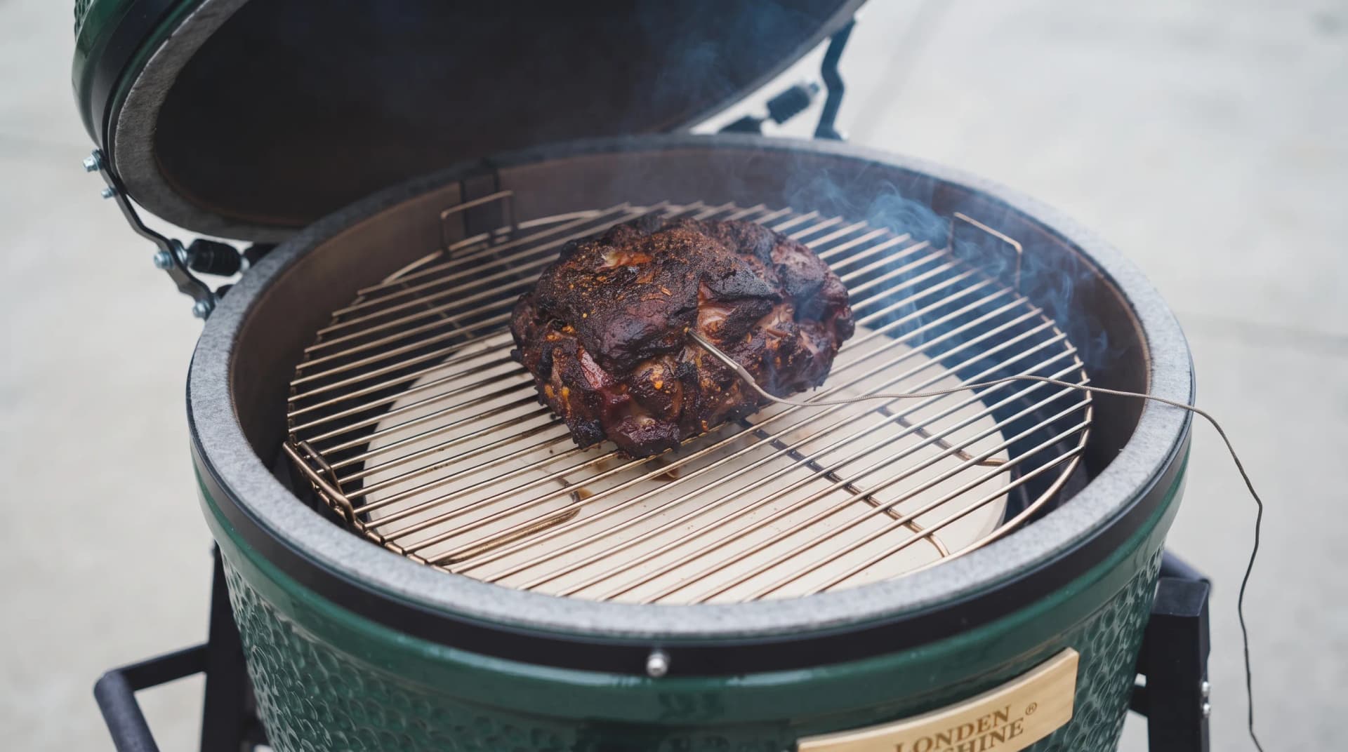 Small pork shoulder resting on the grate of a green ceramic kamado with a probe wire running out