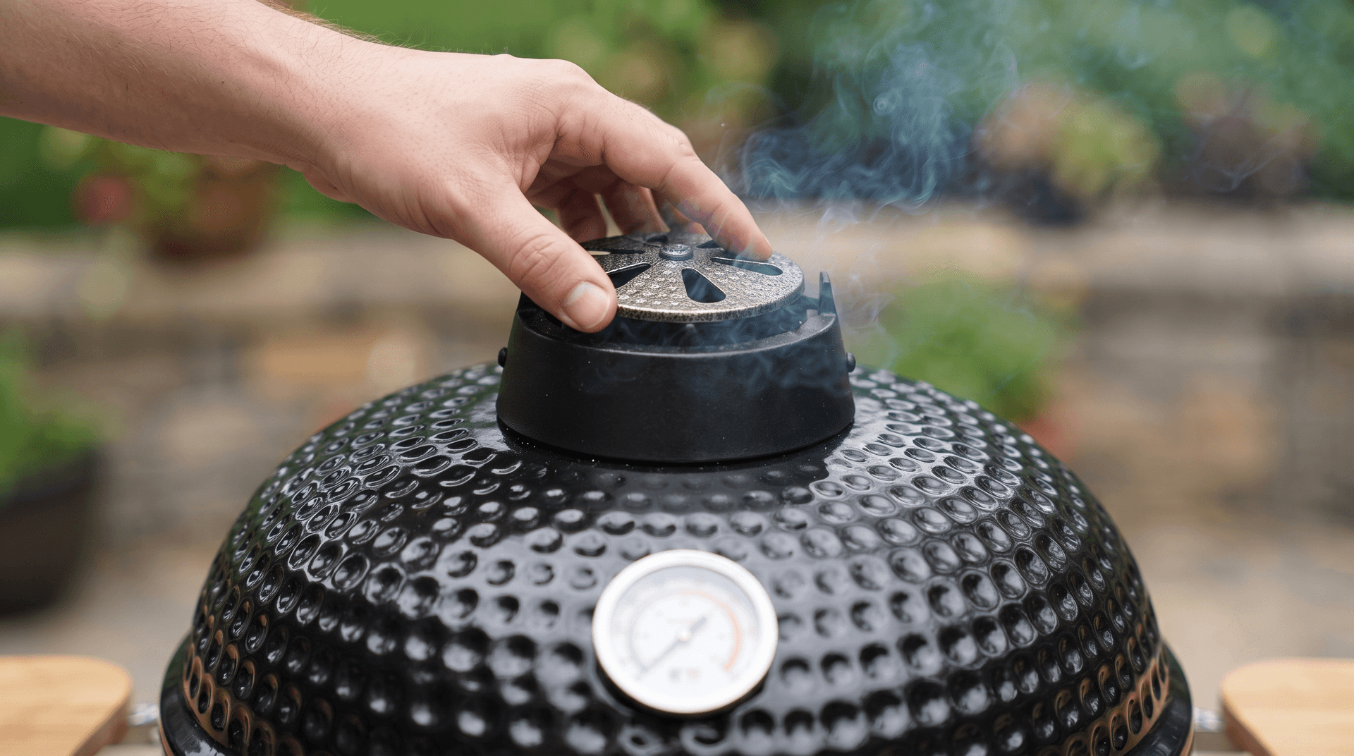 Hand adjusting the top vent on a black ceramic kamado dome with smoke rising